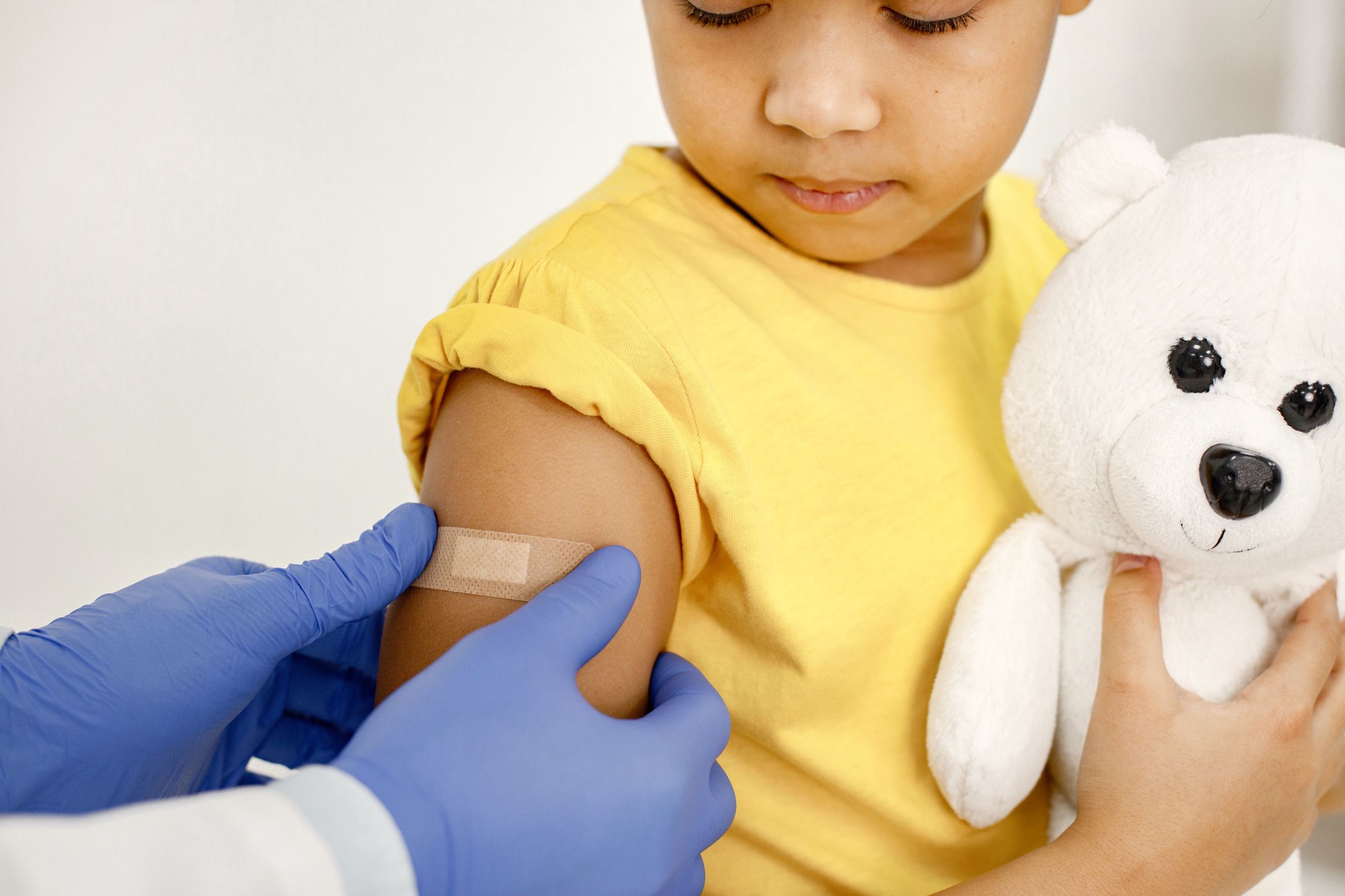 A gloved clinician placing a small plaster on a child's arm as she holds a teddy bear